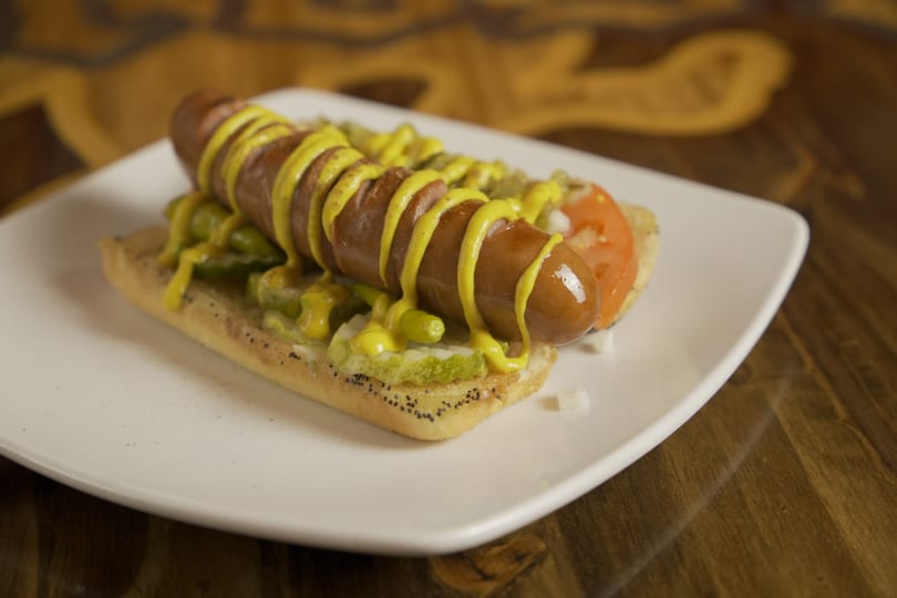Grilled hot dog with mustard, pickles, and tomato on a poppy seed bun.